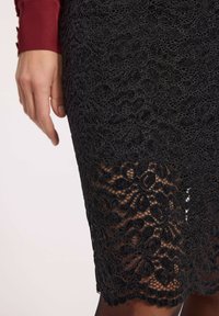 Dłoń spoczywająca obok czarnej spódnicy z koronkowym wzorem, odsłaniająca przezroczystą koronkę na skórzanym podkładzie.