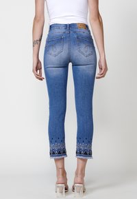 Jeans cropped en denim bleu avec des motifs en diamants noir et blanc brodés à l'ourlet. Comprend des poches arrière et une petite étiquette en cuir.