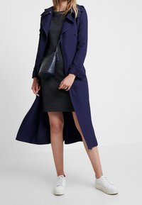 Merihenkinen trench coat, jossa on kaksinkertaiset napit, vyökoriste ja sivuhalkiot, päällä tummassa tekstuurissa oleva mekko ja valkoiset tennarit.