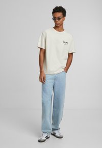 T-shirt en coton beige à manches courtes avec un détail de logo, associé à un jean large bleu clair et des baskets noir et blanc.