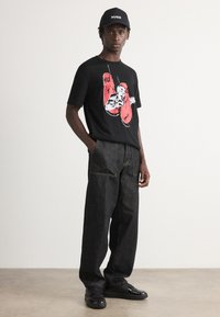 Tricou negru cu un grafic al unor mănuși de box roșii, asociat cu pantaloni din denim închisi la culoare și adidași negri. Accesorizat cu o șapcă neagră.