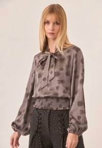 Blusa de seda gris con un lazo en el cuello, que presenta un sutil estampado de hojas y mangas abullonadas, combinada con pantalones de tiro alto negros texturizados.