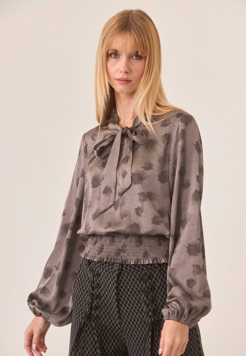 Blusa de seda gris con un lazo en el cuello, que presenta un sutil estampado de hojas y mangas abullonadas, combinada con pantalones de tiro alto negros texturizados.