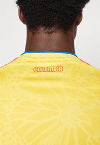 Κίτρινη αθλητική φ�ανέλα με σχεδίαση όπως ιστός αράχνης, μπλε γιακα, και την ένδειξη "ΚΟΛΟΜΒΙΑ" σε κόκκινο χρώμα στο πίσω μέρος.