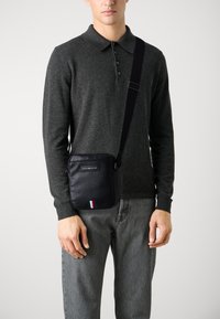 Borsa a tracolla nera con materiale testurizzato, presenta una forma rettangolare e una striscia rossa, bianca e blu nella parte inferiore.