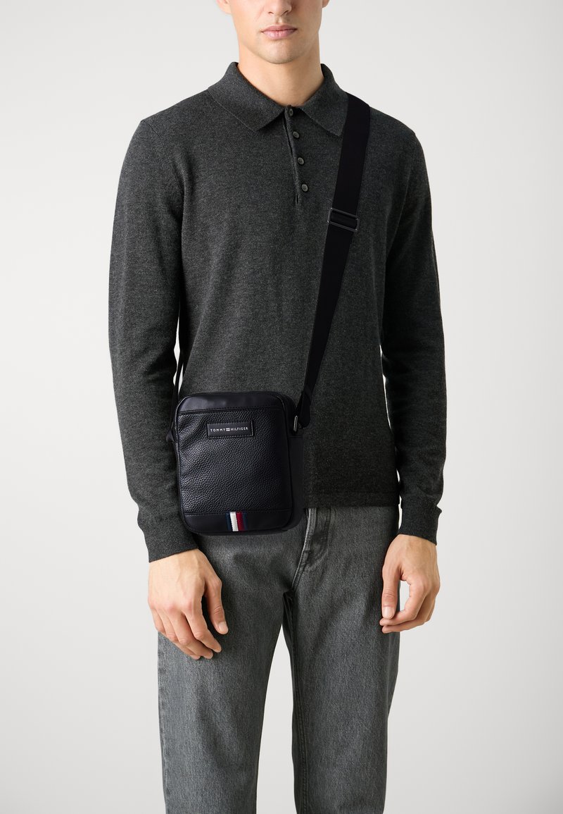 Borsa a tracolla nera con materiale testurizzato, presenta una forma rettangolare e una striscia rossa, bianca e blu nella parte inferiore.