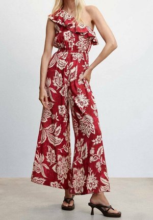 Kobieta ubrana w czerwony kombinezon na jedno ramię z białym kwiatowym wzorem i detalem falbany, w połączeniu z brązowymi sandałami na obcasie, stojąca na szarej podłodze.