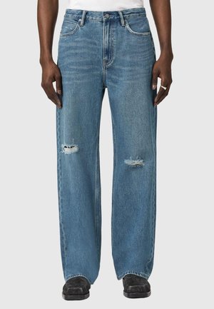Person trägt blau geschnittene Jeans mit abgenutzten Stellen an beiden Knien, kombiniert mit schwarzen Schuhen und einem weißen Hemd.