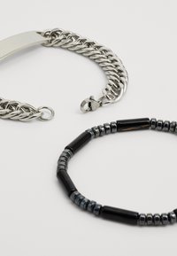 Pulsera de cadena de metal con un acabado plateado pulido y cierre, combinada con una pulsera de cuentas que presenta acentos cilíndricos en negro y gris.
