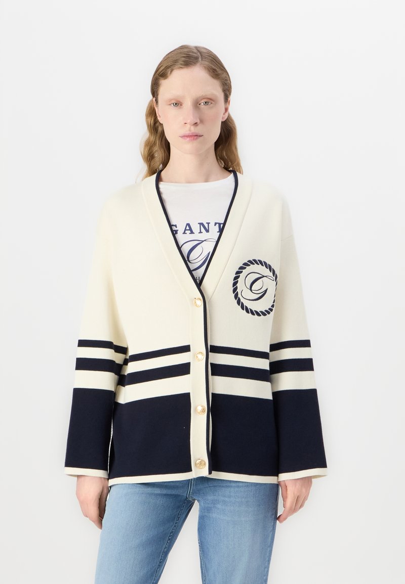 Cardigan crem cu dungi bleumarin, dotat cu un logo pe piept, butoane aurii și mâneci trei sferturi. Material moale și croială lejeră.