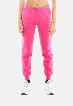 Vue recadrée d’une personne portant un pantalon de jogging rose vif à cordon de serrage, resserré aux chevilles, et des baskets blanches sur un fond uni.