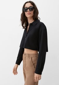 Zwarte cropped button-up blouse met lange mouwen, gecombineerd met bruine high-waisted broeken. De blouse heeft een klassieke kraag en een gladde stoftextuur.