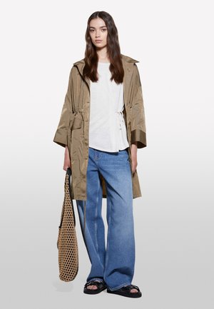 Frau mit langen braunen Haaren trägt eine beige Jacke, weißes Top, weite blaue Jeans, schwarze Sandalen, hält eine beige Netz-Tasche vor hellem Hintergrund.