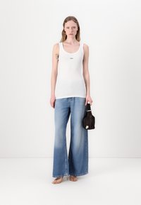 Top sin mangas blanco con textura acanalada, combinado con unos pantalones vaqueros de pierna ancha en un tono azul claro. La modelo sostiene un pequeño bolso de mano marrón oscuro.