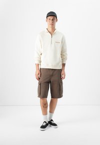 En ung mand står imod en ensfarvet hvid baggrund, iført en cremegul zip-up sweatshirt, brune cargo shorts, sorte sko og hvide sokker.
