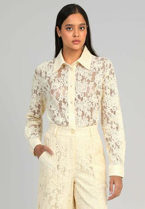 Femme aux longs cheveux noirs, portant une chemise à boutons en dentelle couleur crème et un pantalon taille haute assorti en dentelle, debout avec une main dans la poche.