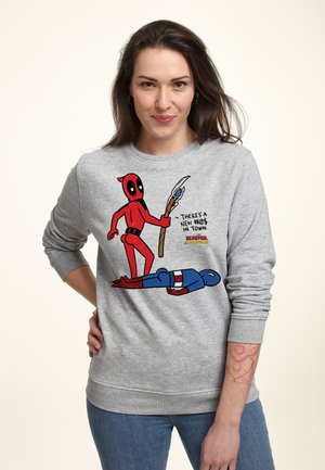 Kvinde iført en grå sweatshirt med en tegneserie af Deadpool, der står over Captain America, holder en stav og tekst, der siger "Der er en ny #@! i byen."