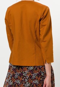 Mujer vistiendo una chaqueta de manga larga de color óxido, con una pequeña abertura lateral, combinada con una falda de estampado floral en negro, blanco, naranja y verde.