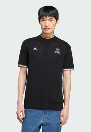 Mann trägt schwarzes kurzärmeliges Polo mit Mercedes AMG Petronas und Adidas Logos, rot-grün-weiße Besätze am Kragen und an den Ärmeln, blaue Jeans.