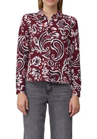 Vrouw in een langärmige blouse met knopen in een rood, wit en zwart paisley bloemenpatroon, gecombineerd met vervaagde zwarte jeans.