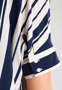 Blouse rayée marine et crème, avec manches courtes à revers et deux boutons blancs ; fabriquée en tissu léger au toucher lisse.