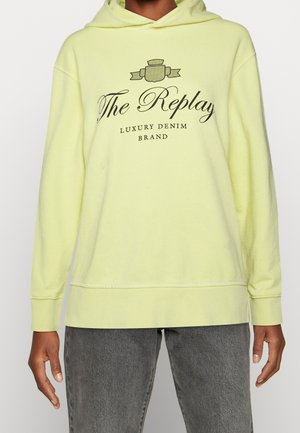 Osoba ubrana w bladożółtą bluzę z kapturem z czarnym napisem "The Replay Luxury Denim Brand" oraz szare dżinsy, ręce swobodnie wzdłuż ciała, neutralne tło.