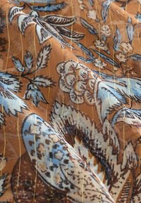Hnedá látka s kvetinovými a vtáčími vzormi v modrej a krémovej farbe, s vertikálnymi zlatými pruhmi, ktoré vytvárajú textúrovaný vzhľad.