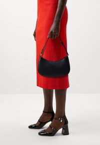 Svart läderhandväska med en böjd form och kort handtag, i kombination med en ljusröd texturerad klänning och lacksvarta klackskor med blockklack.