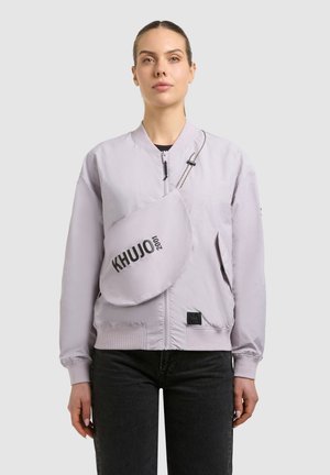 Junge Frau in einer hellvioletten Bomberjacke und mit einer Umhängetasche mit der Aufschrift "KHUJO 2001", trägt schwarze Hosen vor neutralem Hintergrund.