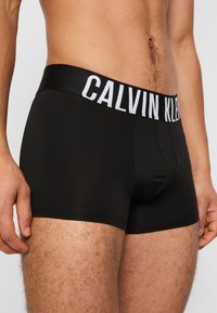 Schwarze Boxer-Brief mit einem dicken elastischen Bund, auf dem "CALVIN KLEIN" in Weiß steht. Hergestellt aus glattem, dehnbarem Material.