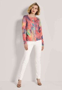 Frau, die einen bunten Knopf-Kardigan, weiße Hose und schwarze, offene High Heels trägt, steht vor einem hellen, neutralen Hintergrund.