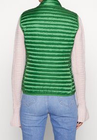 Person iført en grøn quiltet vest over en lys pink strikket sweater med flagrende ærmer og lyseblå denimjeans, vist fra ryggen.
