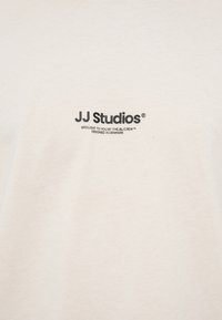 Beige cotton t-shirt featuring "JJ Studios" logo and text printed in dark gray. Simple, soft texture with a crew neck design.