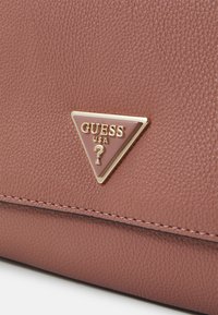 Roza teksturirana umetna usnjena torba z zlatim trikotnim logotipom, ki pravi "Guess USA". Ima čiste šive in gladko površino.