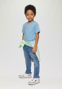 Lichtblauw T-shirt met oranje tekst, groene sweatshirt om de taille geknoopt, blauwe spijkerbroek en witte sneakers met grijze accenten.