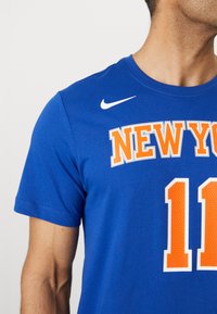 Tricou din bumbac albastru cu inscripția "NEW YORK" scrisă cu litere mari portocalii și numărul "11" în portocaliu pe un fundal alb. Logo-ul Nike pe umăr.