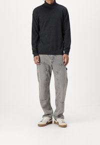 Persona che indossa un maglione a collo alto grigio scuro, pantaloni grigi chiari in stile cargo e sneakers bianche con suole color gomma, in piedi contro uno sfondo bianco.