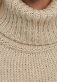 Beige neulottu villapaita, jossa on korkea poolokaulus, ribbineulos ja paksu lankakuosi. Pehmeä ja lämmin ilme.