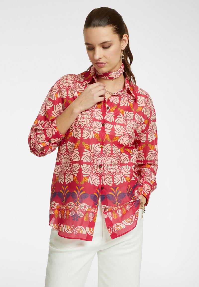 Blusa rossa con motivo a fiori bianchi, dotata di collo, chiusura con bottoni davanti e sciarpa abbinata, indossata con pantaloni bianchi.