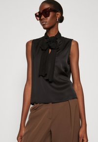 Blusa nera senza maniche con un scollo a fiocco e dettagli plissettati, realizzata in un tessuto liscio. Abbinata a pantaloni marroni.