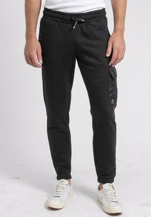 Homme portant un pantalon de jogging noir avec poche latérale et des baskets blanches, debout devant un fond clair uni.