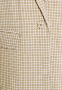 Tissu vichy beige montrant un motif texturé, avec des carreaux blancs. Les caractéristiques incluent un bouton unique et un détail de poche latérale.