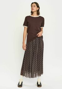 Brun T-shirt med cremefarvet kant, parret med en lang, plisseret brun nederdel med hvide prikker. Svarte sko og hvide sokker fuldender looket.