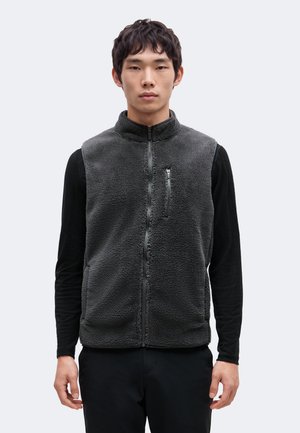 Un homme se tient devant un fond clair uni et présente un gilet polaire gris anthracite sur un haut noir à manches longues et un pantalon noir.