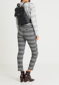 Persona llevando una mochila de cuero negra, pantalones a cuadros grises y negros, una camiseta de manga larga con patrón gris y botines de tacón burgundy.