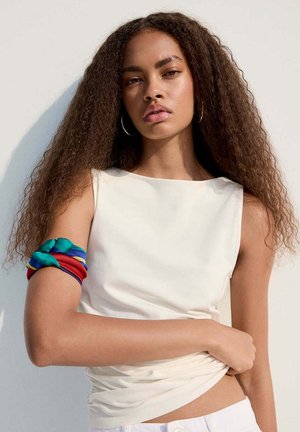 Femme aux longs cheveux bouclés portant un haut blanc sans manches et un tissu coloré enroulé autour de son bras supérieur sur un fond clair.