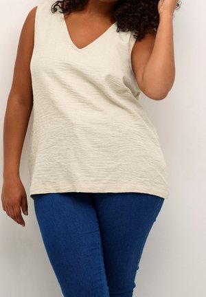 Kobieta w beżowej, bezrękawnikowej bluzce z dekoltem w szpic i dopasowanych niebieskich dżinsach, stojąca na tle gładkiego, jasnego tła.