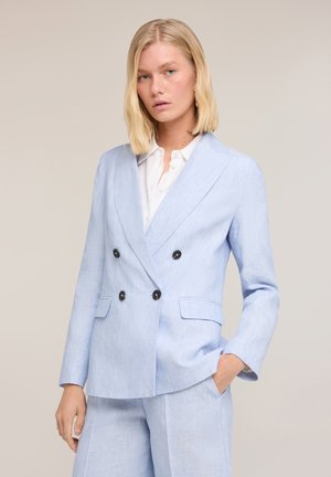 Lyshåret kvinde iført lyseblå dobbeltradet blazer og matchende bukser, hvid skjorte med krave under, den ene hånd i lommen.