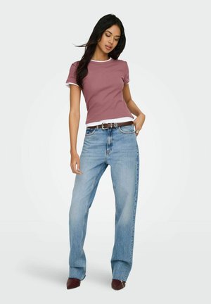 Mujer de pie, vistiendo una camiseta de manga corta color malva, jeans de talle alto azul claro con cinturón marrón y zapatos marrones puntiagudos sobre fondo blanco.