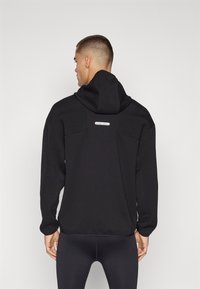 Mustapaita, jossa on teksturoitu paneeli hartioilla, hienovarainen logo takana ja napakka istuvuus. Sileä kangas ja huppu.